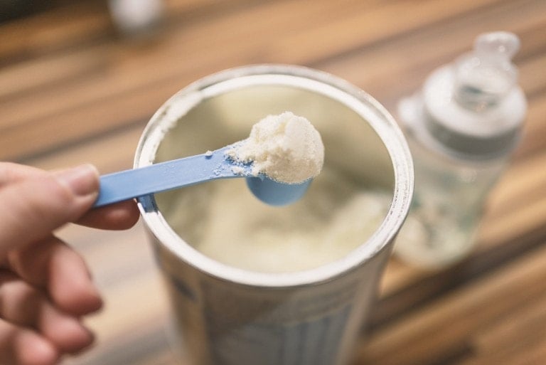 A close-up image of a container of baby formula in powdered form. A hand is holding a blue scoop with a portion of the powder above the open container, ready for preparation. In the background, a baby bottle rests on a wooden surface. Perfect for busy parents listening to their favorite podcast while prepping meals.
