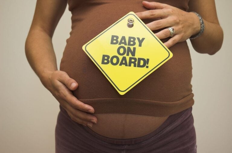 A person wearing a brown shirt holds a yellow sign with black text reading "BABY ON BOARD!" against their pregnant belly, showcasing the body changes during pregnancy. Both hands are cradling the baby bump. The background is plain and light-colored.