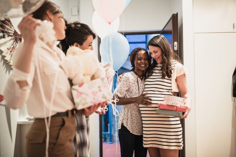 A group of women standing at an indoor entrance, some holding gifts and balloons. One woman in a striped dress is visibly pregnant and is being hugged by another woman. The setting appears festive, likely a baby shower to celebrate the new mom.