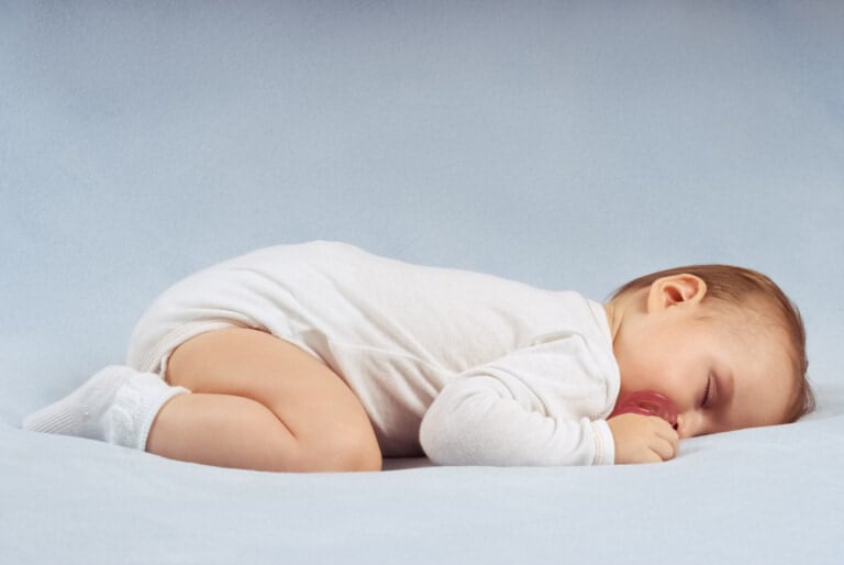 A baby lies on its side on a light blue surface, wearing a white onesie and socks, holding a pacifier in their mouth. The baby appears to be sleeping peacefully, which can help reduce the risk of SIDS.