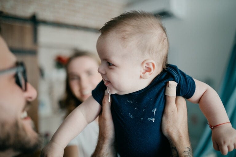 A baby in a blue onesie is being lifted into the air by an adult. The baby has some drool on their chin, possibly from a recent spit-up. In the background, a smiling man with glasses and a blurred woman are visible. The focus is on the smiling baby.