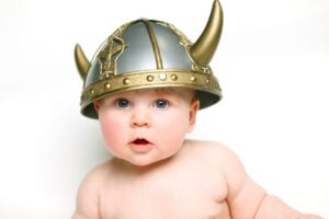 A baby with blue eyes and a slightly open mouth is wearing a silver and gold Viking helmet with horns, reminiscent of ancient Norse names. The baby is shirtless and sitting against a plain, white background.