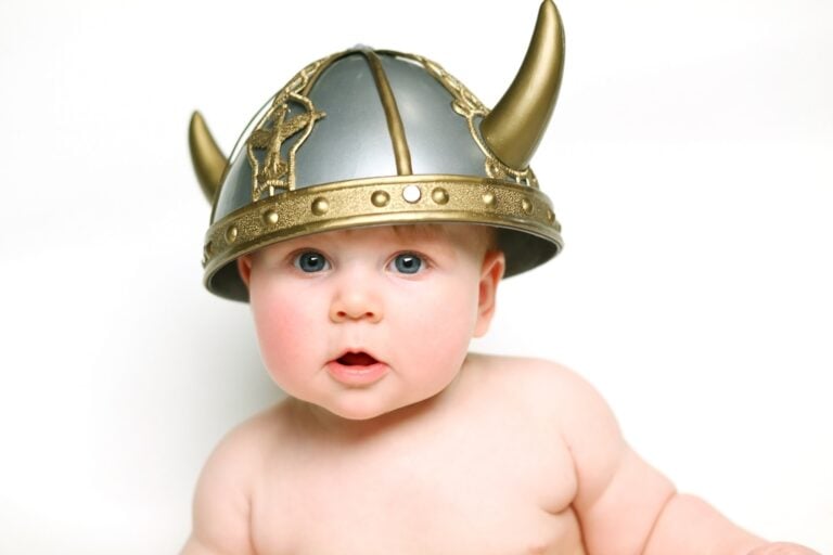 A baby with blue eyes and a slightly open mouth is wearing a silver and gold Viking helmet with horns, reminiscent of ancient Norse names. The baby is shirtless and sitting against a plain, white background.