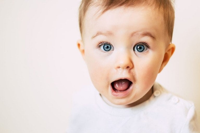 A baby with striking blue eyes and short brown hair is looking directly at the camera with a surprised expression and mouth open. The baby is wearing a white outfit, set against a plain white background.