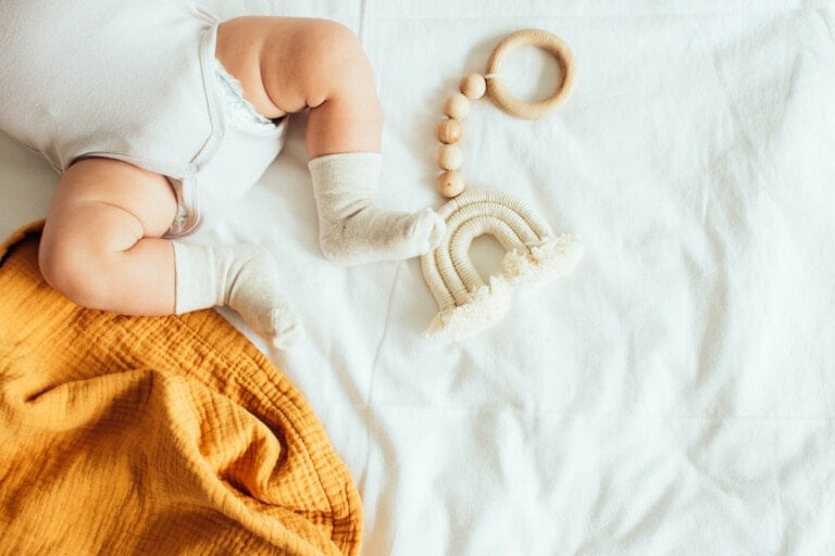 A baby wearing a light-colored onesie and socks lies on a white sheet next to a yellow blanket and a wooden beaded baby toy shaped like a rainbow. Surrounded by natural baby products, the baby's upper body is not visible in the image.