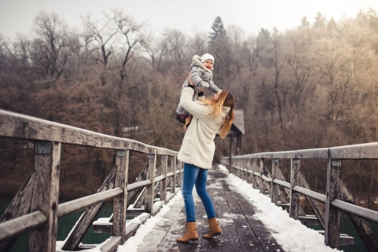 A person finds joy lifting a small child into the air while standing on a wooden bridge. Both are dressed in warm, winter clothing. The bridge is surrounded by a forested area with bare trees, and some snow is visible on the bridge's surface and around the area, capturing a perfect moment of winter time delight.