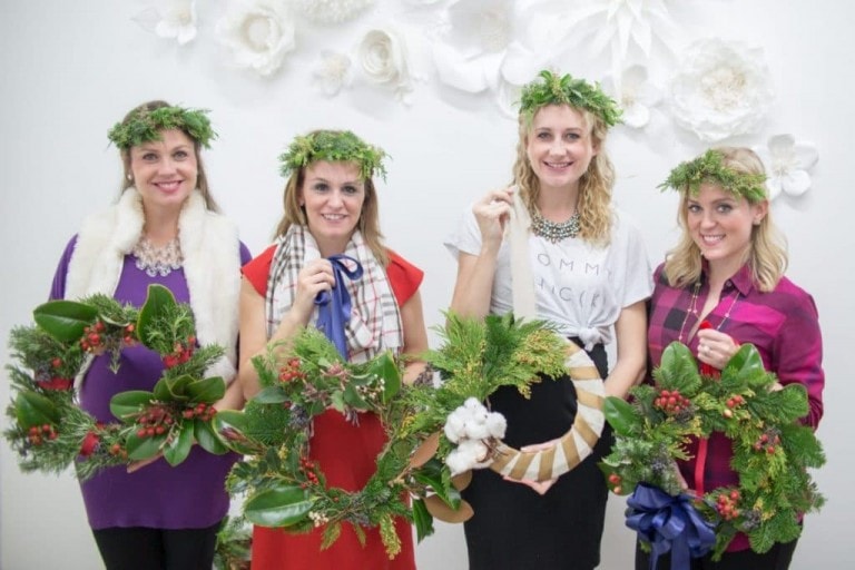 Four women stand in front of a white wall adorned with floral decorations. They are each holding wreaths made of greenery and berries, showcasing their wreath-making skills, and wearing leaf crowns. Dressed in casual to semi-formal attire, they smile warmly at the camera, celebrating their creative endeavor.