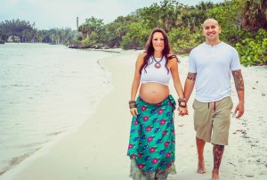 A pregnant woman in a white crop top and green skirt with a floral pattern walks hand-in-hand with a man in a white shirt and beige shorts on a beach, enjoying their babymoon. They are both smiling. Greenery and water are visible in the background.