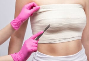 Person wearing a beige tube top is having a pre-surgical consultation. Gloved hands, holding a scalpel, are poised near the top. The scene suggests a medical or cosmetic procedure setting.