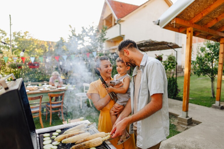 A family of three stands by a grill in a backyard, enjoying some fun family activities. The man is grilling corn, while the woman holds a young child. There are tables with food and decorations in the background, indicating a gathering or celebration. The scene is outdoors with a house and patio nearby.