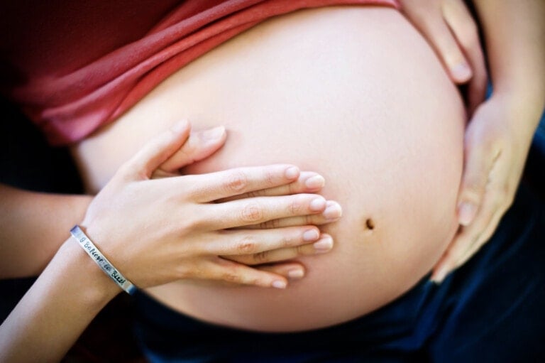 A pregnant person's belly is shown with hands gently resting on it. One hand belongs to the person wearing a pink shirt, while another person's hand, wearing a bracelet that says "DREAM PLAN DO," gently touches the belly — a scene that might make you feel confident you've found the right doula for you.