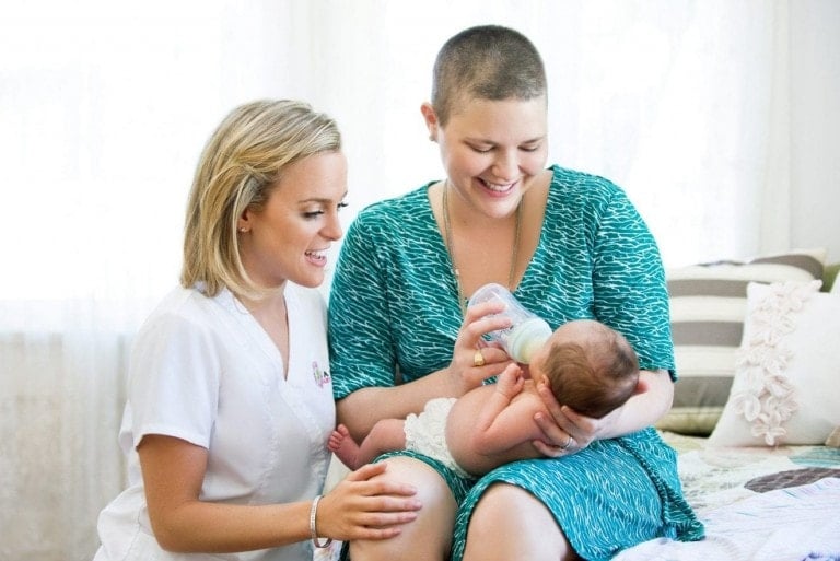 A woman with short hair is bottle-feeding a baby while sitting on a bed. Another woman with shoulder-length blonde hair, dressed in a white uniform, is kneeling beside them, touching the woman's knee and smiling. It’s moments like these that are often shared on postpartum doula podcasts.