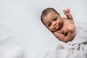 A baby with dark skin is lying on a light-colored textured blanket, partially covered by a soft cloth. The baby is looking at the camera with a small smile and has its arms slightly raised, perfect for parents considering boy names that start with U.