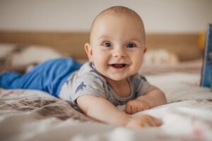 A baby lying on a bed on their stomach, wearing a gray outfit with small prints and blue pants. The baby is smiling, with wide eyes and arms extended forward. If you're looking for inspiration, consider popular boy names that start with G as you gaze at this joyful scene in the blurred indoor setting.