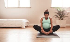 A pregnant woman sits on a yoga mat in a butterfly pose, embracing the benefits of exercise during pregnancy. She wears a turquoise tank top and black leggings in a bright room with wooden floors, a white bed, and a potted plant in the background.