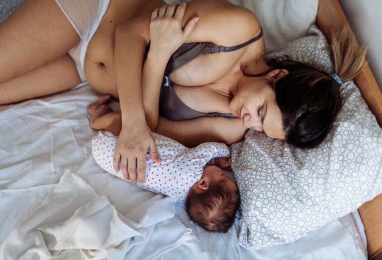 A woman lies on a bed next to a baby, both covered with a white sheet. The woman, experiencing lochia, is wearing a bra and underwear and gently touching the baby, who is dressed in a white onesie with red polka dots. They appear to be resting in a calm and intimate moment.