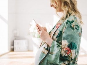 A pregnant woman stands indoors holding a smartphone, presumably browsing the best pregnancy apps. She is wearing a green floral robe and her blonde hair is styled in loose waves. The background is bright and includes a blurred sign and some floral arrangements.