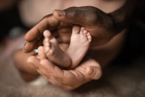 An adult's hands gently cradle a 1-month-old baby's feet. The image focuses closely on the contrast between the large adult hands and the tiny baby feet, emphasizing care and tenderness. The background is blurred, keeping attention on the hands and feet.