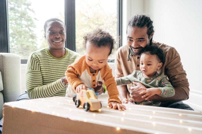 A family of four is gathered indoors. Two adults sit beside each other, smiling and watching two small children. One child plays with a language development toy car on a box, while the other child is held by an adult. The scene appears warm and joyful.