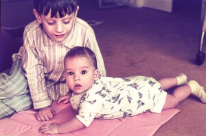 A young child with classic boy names in pajamas is sitting on the floor next to a baby, who is lying on a pink pad. The baby, dressed in a white onesie with a colorful pattern, is looking up with wide eyes. The room has a carpet and some furniture is visible in the background.