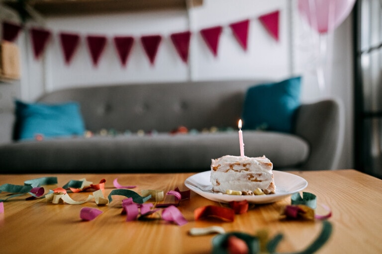 A slice of cake with a single lit candle on top is placed on a plate on a wooden table. In the background, a gray sofa with blue cushions and a string of pink triangular flags hanging on the wall can be seen, along with colorful streamers—perfect decor for a first birthday bash.