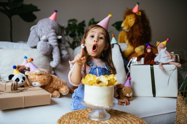 A young child is seated on a couch surrounded by stuffed animals and gift-wrapped presents, reveling in birthday fun. The child is wearing a party hat and appears to be talking or singing, pointing with one finger. A small cake with yellow flowers sits on a woven table in front.