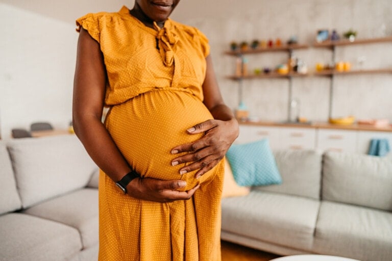 A pregnant person wearing a yellow dress cradles their belly with both hands. They are standing in a modern living room with a grey couch and shelves with decorations in the background, perhaps contemplating the 10 commandments of pregnancy.