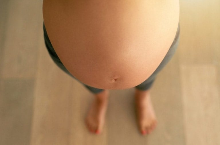 The image shows a close-up top view of a pregnant woman’s belly covered in maternity leggings, with her feet visible in the background. She is standing on a wooden floor, and her toenails are painted red.