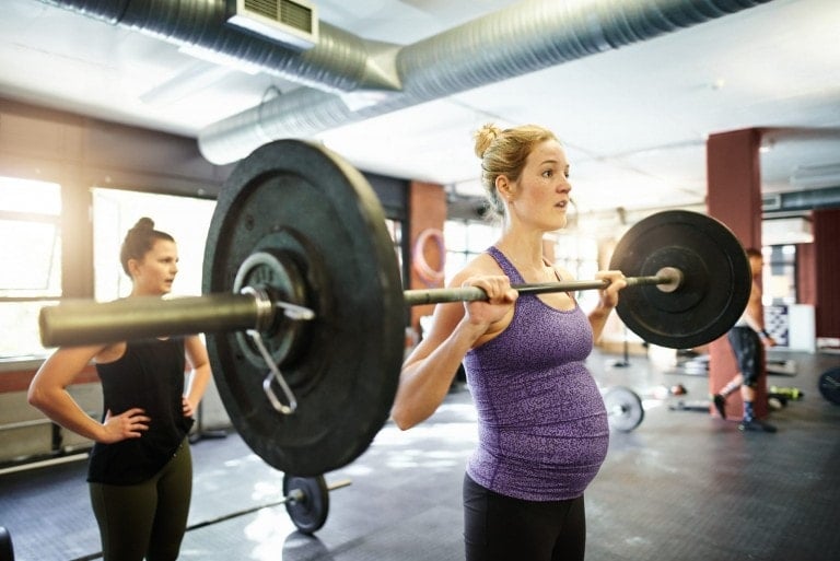 A pregnant woman in a purple tank top is lifting weights, holding a barbell on her shoulders in a gym. Another woman stands behind her with her hands on her hips. The gym, equipped with various workout gear, boasts large windows that flood the space with natural light.