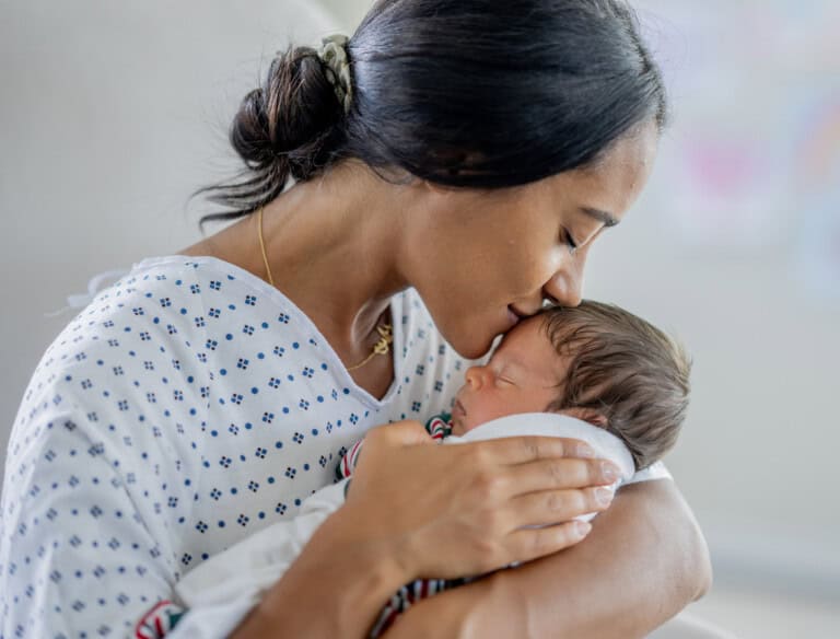 A new mother in a hospital gown gently holds and kisses her newborn baby wrapped in a striped blanket. They are in a bright room with soft lighting.