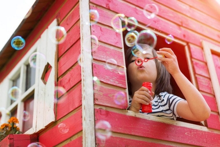 A child with shoulder-length hair, wearing a striped shirt, blows soap bubbles from the window of a red wooden playhouse. The surrounding area is filled with floating bubbles, showcasing the joy of outdoor toys for kids.