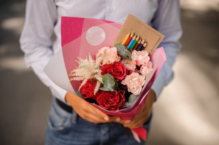 A person holding a bouquet of red and blush pink flowers wrapped in pink paper, with a small pack of colored pencils attached near the top—a perfect teacher gift. The individual is dressed in a light blue, button-up shirt and blue jeans. The background is blurred.