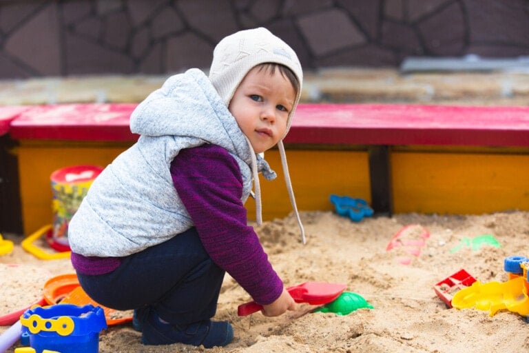 A young child wearing a gray hat and hoodie sits in a sandbox, holding a red toy shovel. Various colorful toys are scattered around in the sand as the background features the red and yellow edge of the sandbox and a stone wall. A demanding toddler nearby tries to grab one of the toys, causing some commotion.