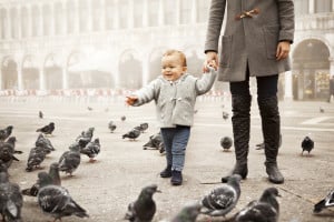 A toddler holding an adult's hand walks among pigeons in a large, open square. The child is dressed in a light gray jacket and blue pants, while the adult wears a dark gray coat and black boots. The background features an arched building with fog, evoking images of charming Italian boy names like Luca or Matteo.