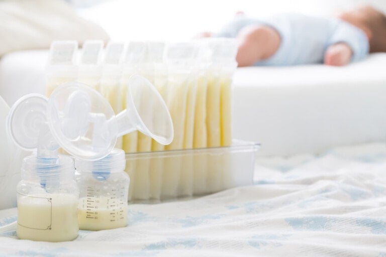 A breast pump and bottles containing milk are in the foreground, essential tools for power pumping. Behind them, several bags of stored breast milk are lined up in a container. In the background, a baby dressed in blue is lying down on a white surface.