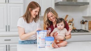 Two women in a kitchen smile while preparing byheart formula. One holds a baby, the other holds formula items. Appliances behind.