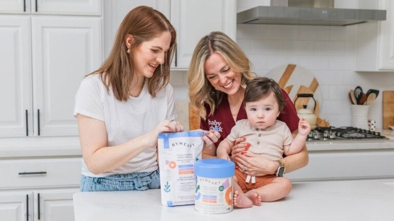 Two women in a kitchen smile while preparing byheart formula. One holds a baby, the other holds formula items. Appliances behind.
