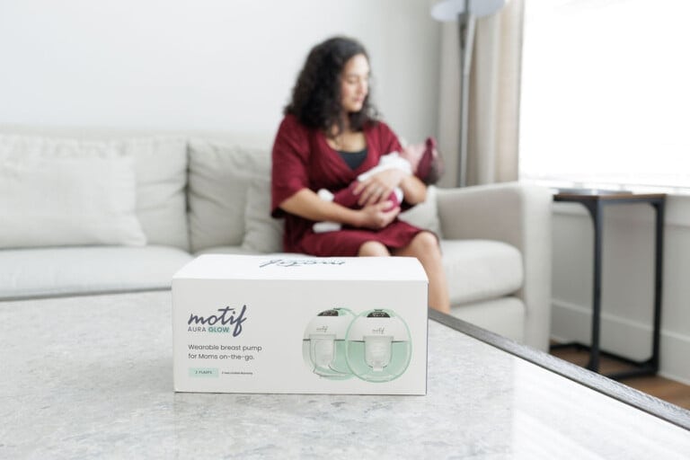 A white box labeled Motif Aura Glow containing a wireless breast pump is placed on a table. In the blurred background, a woman in a red dress is sitting on a sofa, nursing her baby.