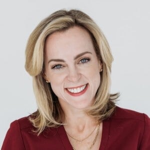 Nina Spears with shoulder-length blonde hair is smiling at the camera. They are wearing a maroon top and a thin gold necklace. The background is plain and light-colored.