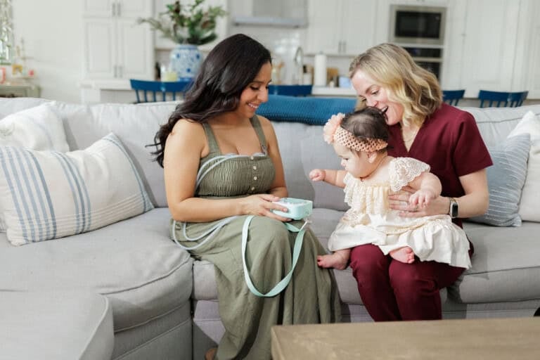 A woman in a green dress and another in a maroon outfit sit on a couch with a baby. The baby, wearing a lace dress and headband, reaches toward the cup held by the woman in green. The living rooms neutral-toned decor serves as a serene motif throughout the setting.