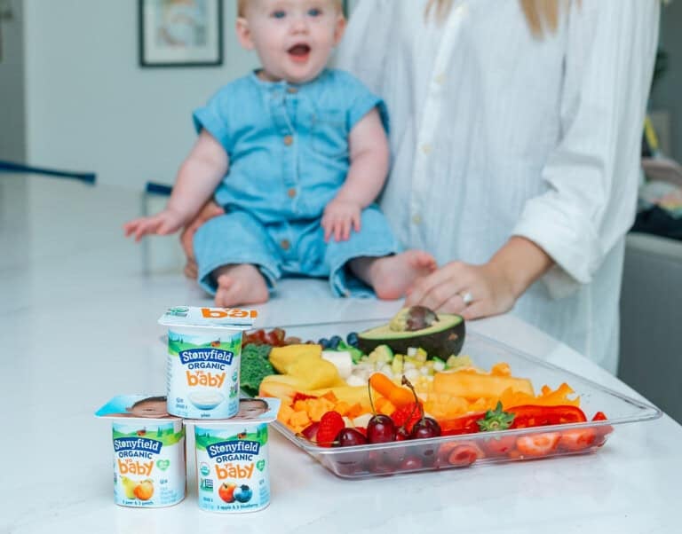 A baby in a blue outfit sits on a counter next to an adult in a white shirt. In front of them, there is a colorful plate filled with assorted fruits and vegetables. Additionally, there are four tubs of Stonyfield Organic Baby yogurt on the side.