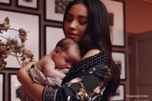 A woman with long dark hair holds a sleeping baby wrapped in a blanket. She is wearing a patterned robe and standing in a room with framed pictures on the wall. The image has a warm tone, capturing an intimate moment. The text "IG @shaymitchell" is visible in the lower right corner, hinting at celebrity baby names.