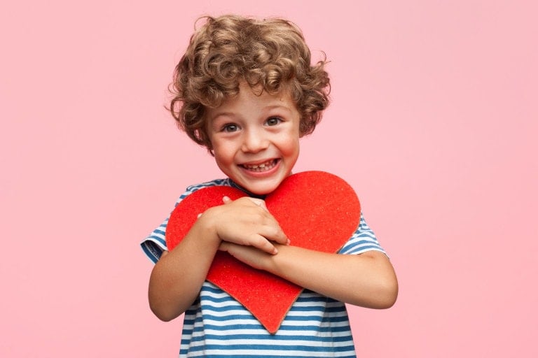 A young child with curly hair and a striped shirt is smiling and holding a large red heart against a pink background, perfect for showcasing Valentine's Day gifts.