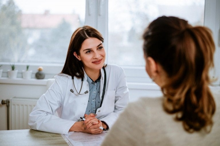 A female doctor, possibly a pediatrician podcast host, is sitting at a desk, attentively listening to a female patient seated across from her. The doctor has a stethoscope around her neck and is looking at the patient with a smile. There are windows and potted plants in the background.