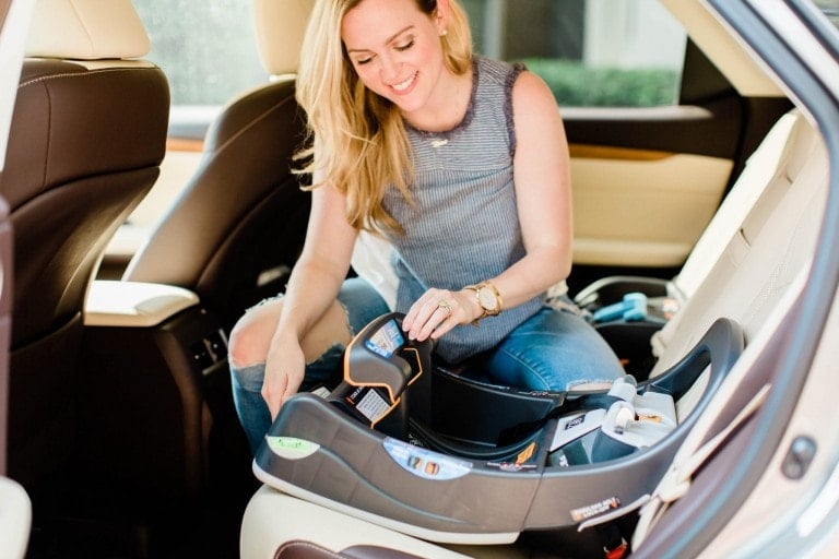 A woman installs a car seat in the rear seat of a car. She is smiling and appears to be adjusting the straps, ensuring car seat safety. The car seat is gray and positioned in the center of the backseat. The interior of the car has beige and brown accents.
