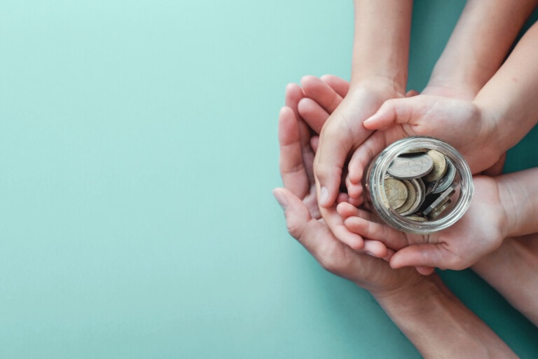 A pair of adult hands and a pair of children's hands hold a clear jar filled with coins against a light blue background. The cupped hands symbolize protection or sharing, reflecting valuable resources for parents.