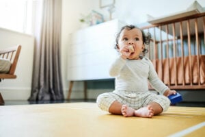 A 10-month-old baby wearing a grey shirt and plaid pants sits on a yellow mat in a room with a crib, dresser, and chair. The child is holding a blue object in one hand and has the other hand near their mouth. The room is well-lit with natural light coming from a window, reflecting key 10-month-old milestones.