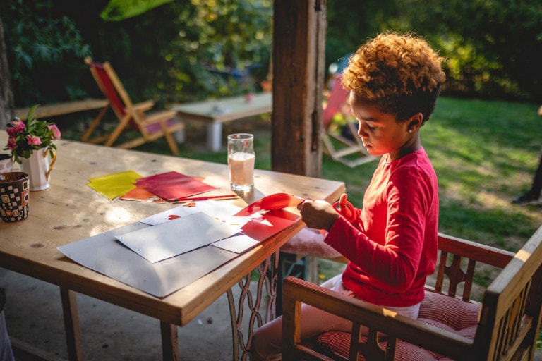 A child in a red long-sleeve shirt sits at an outdoor table, engaging in crafts for kids by cutting red paper with scissors. The table is scattered with craft supplies, including colorful paper and a glass of juice. In the background, greenery surrounds a wooden chair.