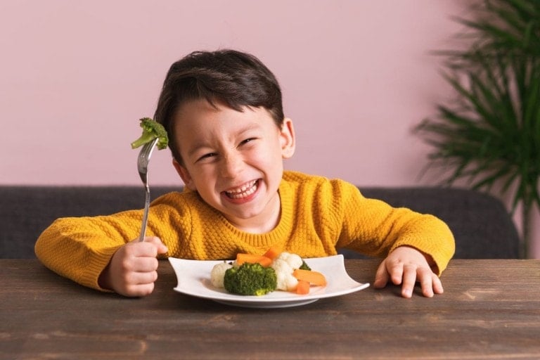 A smiling child in a yellow sweater sits at a wooden table in front of a plate of kid-friendly vegetables. The child is holding a fork with a piece of broccoli on it. There is a pink wall and a green plant in the background.
