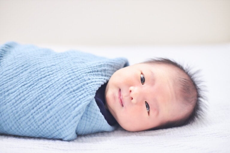 A newborn baby is lying on a white blanket, wrapped in a light blue textured blanket. The baby has short, dark hair and is looking toward the camera with a calm expression. The background is plain and softly lit, debunking any baby myths about first-time photo perfection.